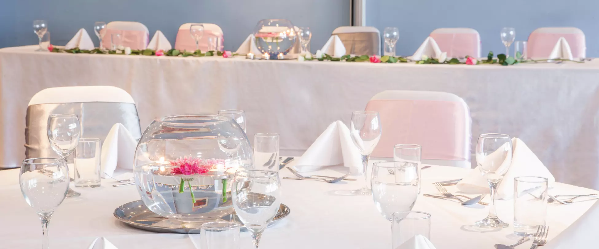 Tables set up for a wedding breakfast with white table and chair covers, pink ribbons and glass centrepieces..
