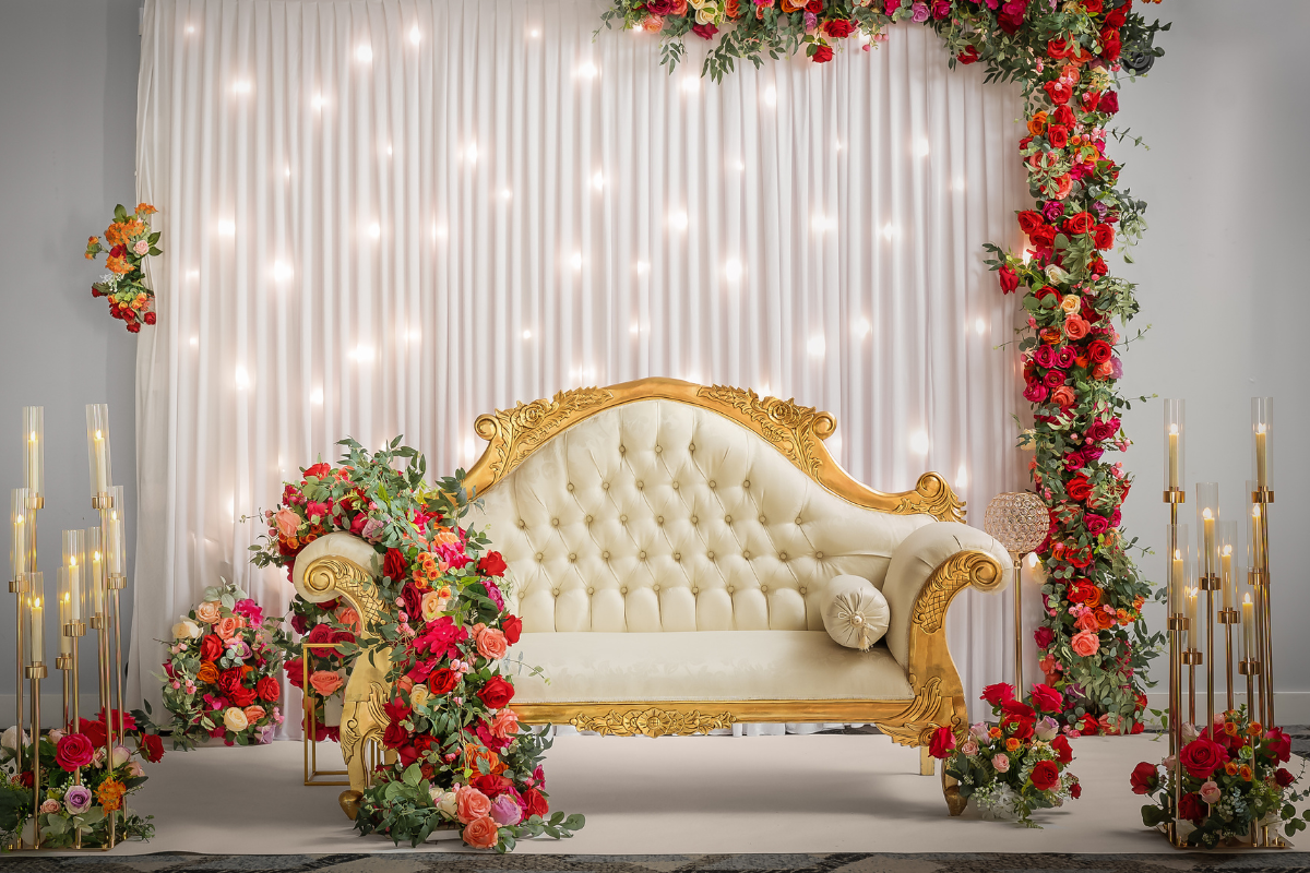 A cream and gold chair sits in front of a lit backdrop. Red, orange, and pink flowers are draped over one arm of the sofa and over the top and right side of the backdrop. There are white candles in gold stands to either side and even more flowers..